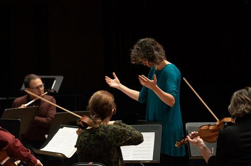 A music conductor stands in front of the ensemble with her hands outstretched, no baton. The back of a viola player in dark green ad a flute player in dark red is visible.