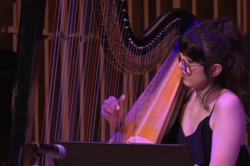 In a pink hue light, Samantha Murray has her right hand plucking strings of a grand harp against a dark wooden backdrop.