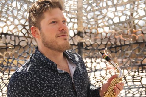 Nick stands holding a tenor saxophone, and is in front of a tightly patterned sculpture.