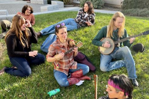 Members of the bluegrass ensemble sit with their instruments (banjos, violins, guitars) on the lawn. The grass is very green and it is a very sunny day.